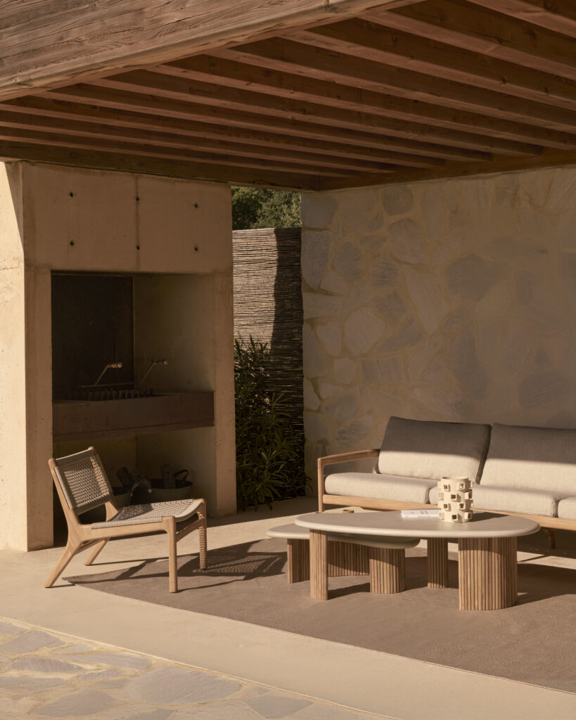 Terrasse extérieure comprenant une chaise longue d'extérieur Jack Woven, un canapé rembourré et une table basse, avec un mur de pierre et un plafond en bois partiellement visibles en arrière-plan.