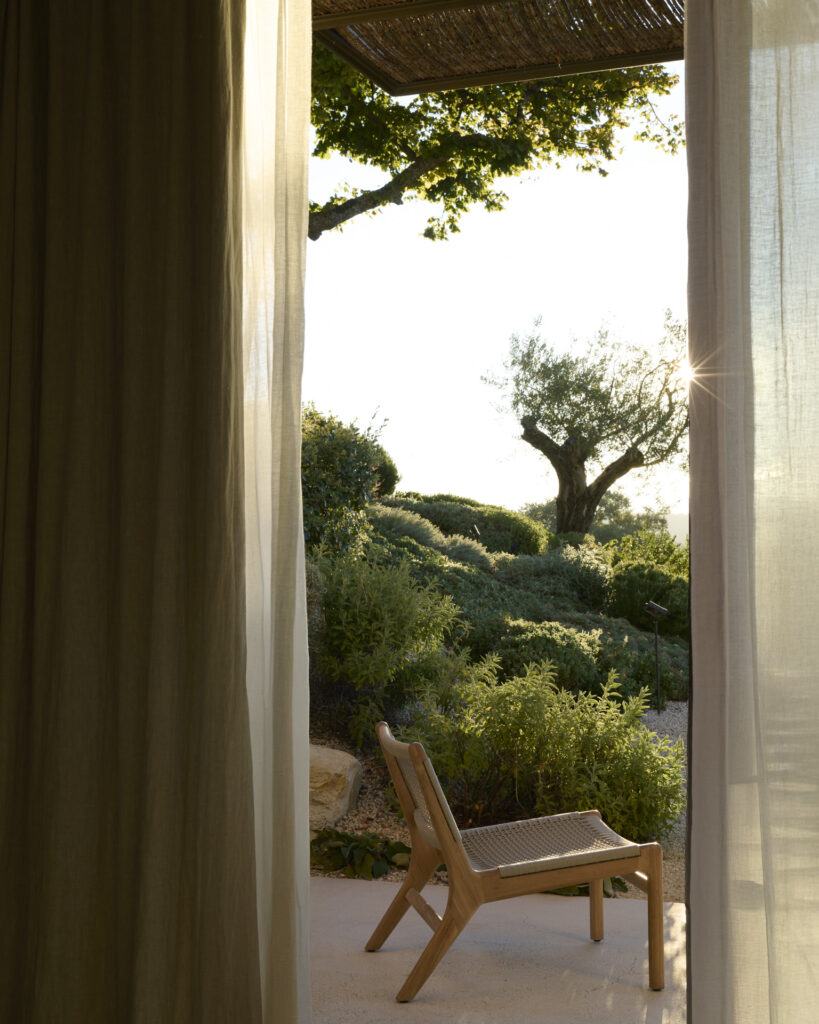 Une chaise longue d'extérieur Jack Woven est posée sur une terrasse, face à un jardin luxuriant avec un olivier, et est visible à travers des rideaux transparents.