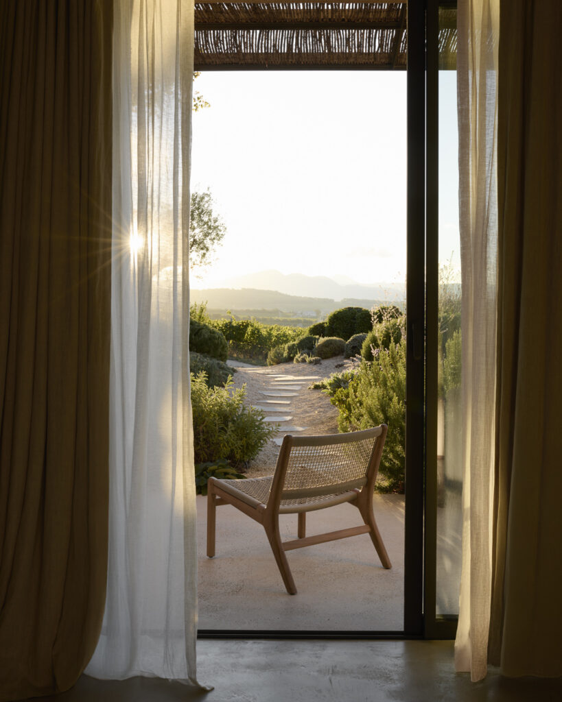 Une chaise longue d'extérieur Jack Woven est installée sur une terrasse, surplombant les montagnes et la verdure à travers une porte coulissante en verre encadrée de rideaux.