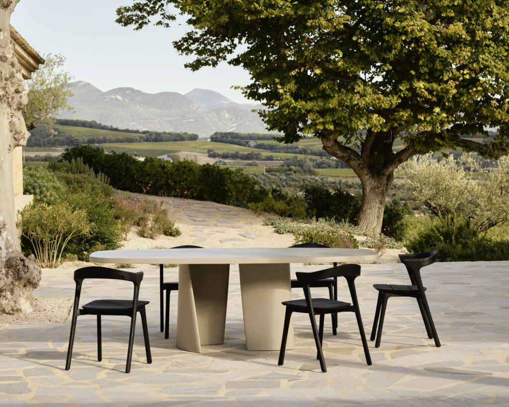 Une scène de repas en plein air accueillante présente une table à manger ovale Canyon avec quatre chaises noires sur une terrasse en pierre. Un arbre majestueux et des collines vallonnées rehaussent la toile de fond sous un ciel clair, créant une expérience idyllique en plein air.