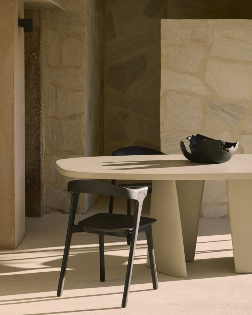 Une chaise en bois et une table à manger d'extérieur ovale Canyon avec un bol décoratif sombre sont placées dans un coin ensoleillé contre un mur à la texture de pierre.