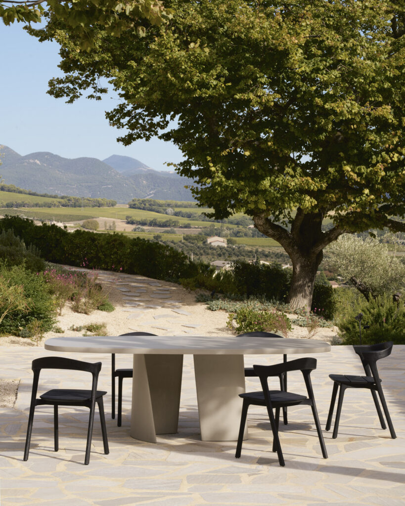 Une scène de repas en plein air accueillante met en valeur une table à manger ovale d'extérieur Canyon entourée de cinq chaises noires élégantes sur une surface en pierre, avec des arbres et des montagnes majestueuses en toile de fond époustouflante.