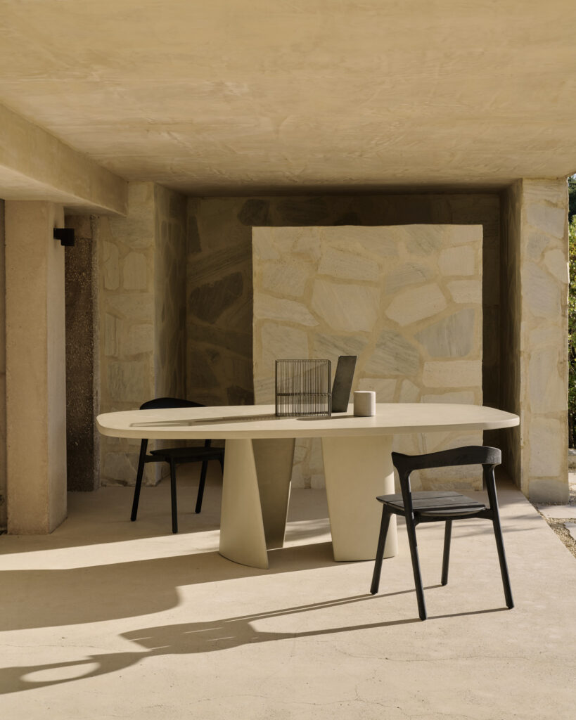 Un patio extérieur minimaliste avec un mur de pierre met en valeur la table à manger d'extérieur ovale Canyon en beige moderne et deux chaises noires sous un plafond en béton, évoquant une atmosphère de canyon sereine.