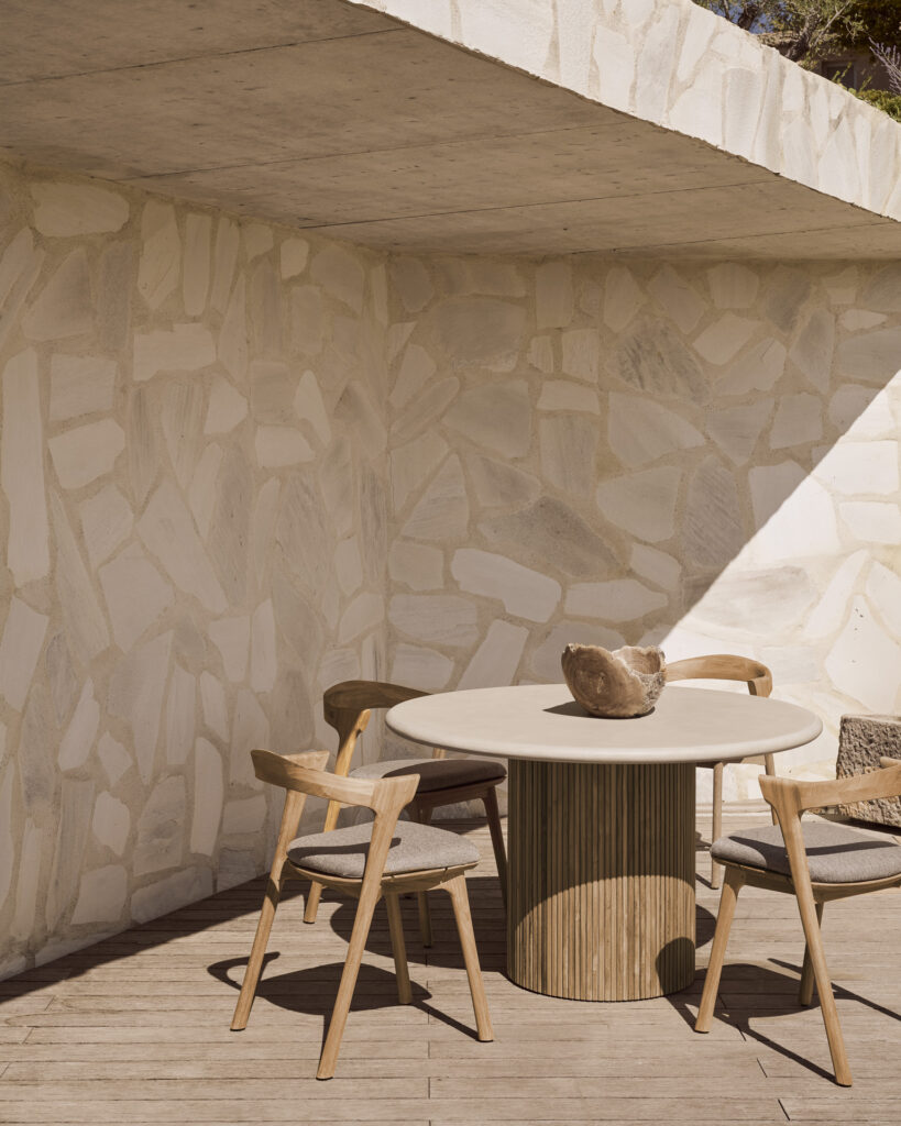 Une terrasse extérieure avec la table à manger Roller Max, trois chaises en bois et un bol décoratif. Un mur en pierre et un toit partiel servent de toile de fond tandis que la lumière naturelle projette des ombres géométriques, créant une atmosphère accueillante.