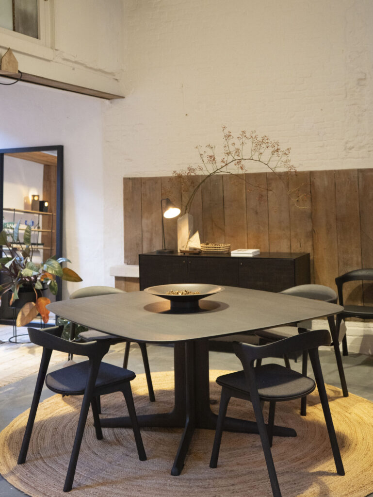 Une salle à manger moderne est dotée de la table carrée Expo Table Corto avec quatre chaises noires et une pièce maîtresse élégante. Des panneaux muraux en bois et un buffet décoré rehaussent l'arrière-plan, tandis qu'un tapis tissé ajoute de la chaleur au sol.