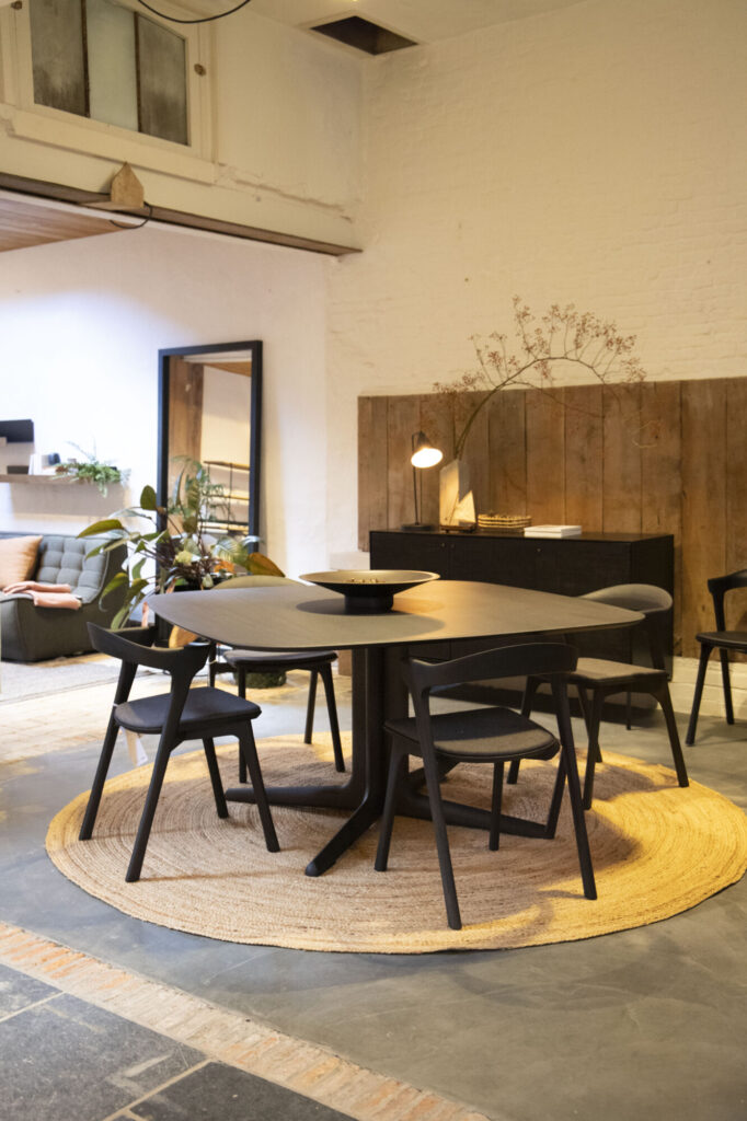 Une salle à manger moderne présente une élégante table noire Expo Corto avec des chaises sur un tapis rond. Un canapé confortable et des plantes luxuriantes rehaussent l'ambiance, tandis qu'un panneau en bois et une lampe fournissent une lumière chaleureuse venant de la droite.