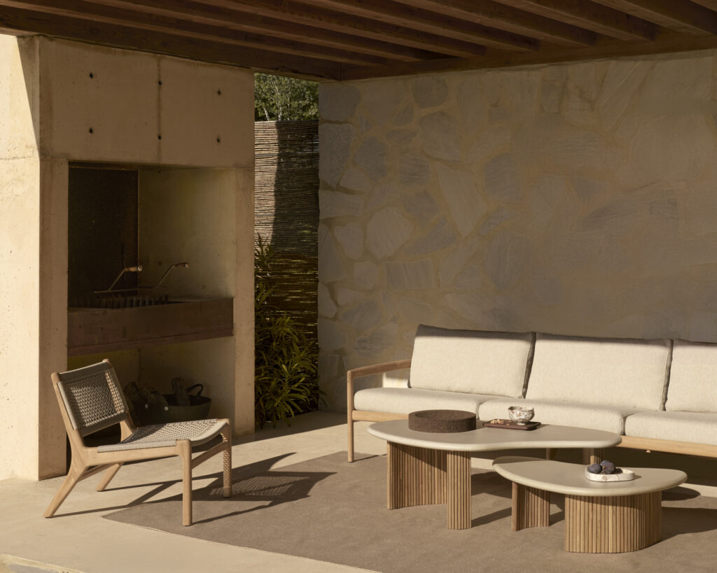Espace salon extérieur avec une chaise en bois, un canapé rembourré et deux tables basses de jardin. Un mur en pierre en arrière-plan et une pergola couverte au-dessus offrent le mélange parfait de décor minimaliste. Cet élégant mobilier extérieur invite à la détente et au confort.
