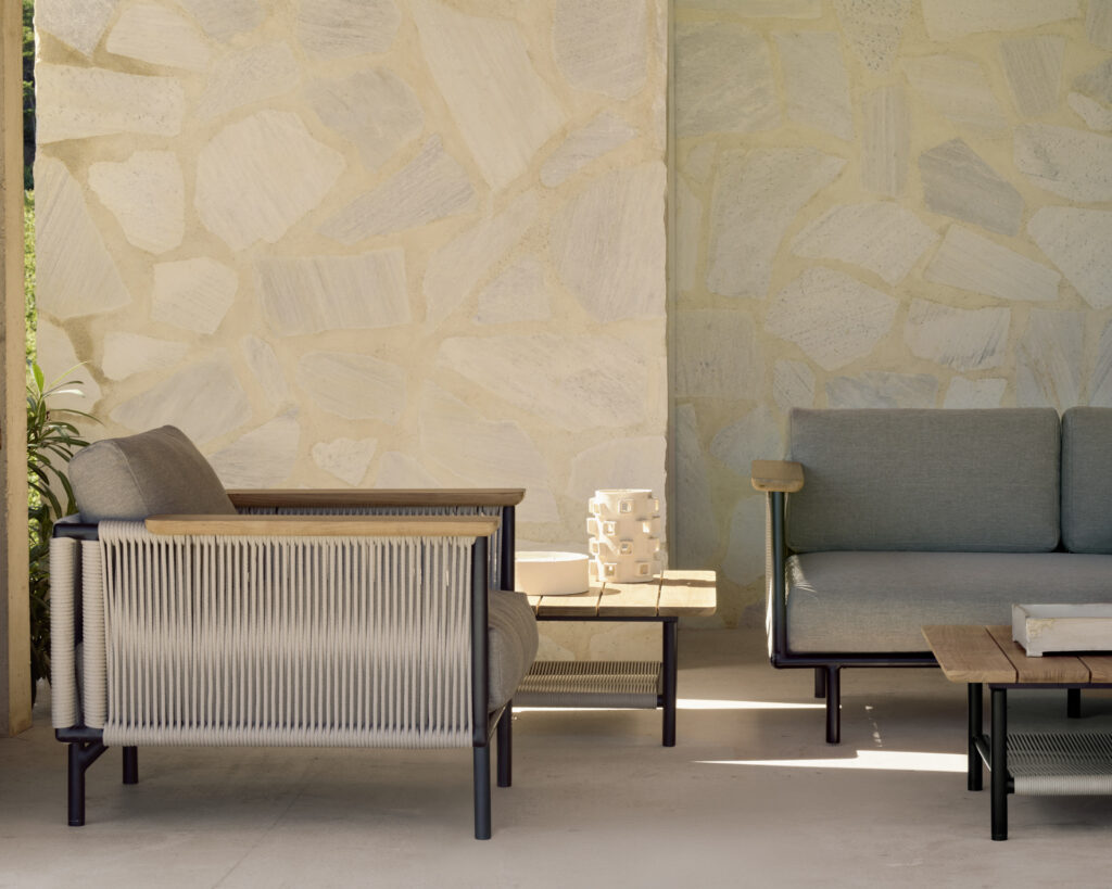 Espace salon extérieur avec un fauteuil couleur crème recouvert d'un luxueux tissu huître et un canapé vert sauge sur une terrasse en pierre carrelée. Une petite table avec des livres est placée devant le canapé. Un mur en pierre de Corbey forme l'arrière-plan, créant une oasis accueillante pour la détente.