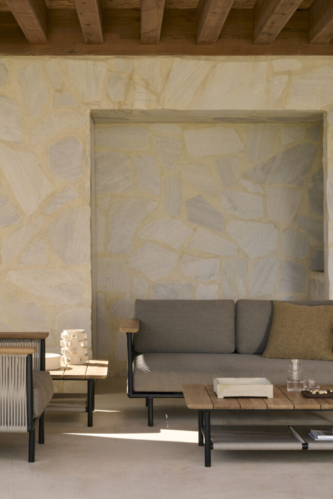 La terrasse extérieure est dotée d'un mur en pierre beige, d'un canapé d'extérieur Corbey, d'une table basse en bois et d'une petite table d'appoint en bois avec une pièce décorative. Un toit en bois ombrage élégamment la zone.