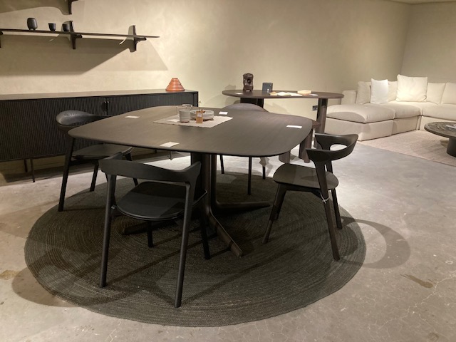 Une salle à manger moderne avec l'Expo - Table Corto en bois foncé, quatre chaises noires et un tapis rond dans une pièce minimaliste avec un canapé, un buffet et des étagères - parfait pour des réunions élégantes.