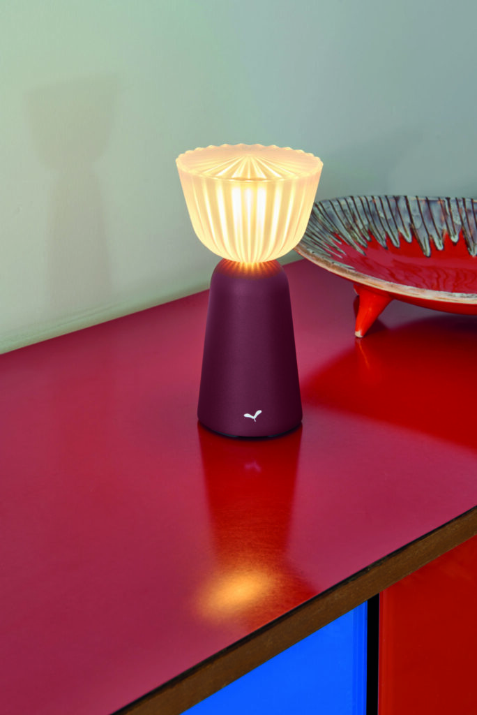 La Lampe Swiing 7, avec son dessus texturé et lumineux distinctif, orne une surface rouge à côté d'un bol décoratif.