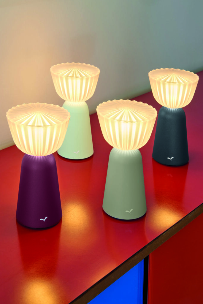 Quatre lampes de table Lampe Swiing 7, aux motifs en forme de fleurs, ornent une surface rouge. Leurs sommets lumineux illuminent les bases dans des tons modernes de violet, vert menthe, turquoise et bleu foncé.