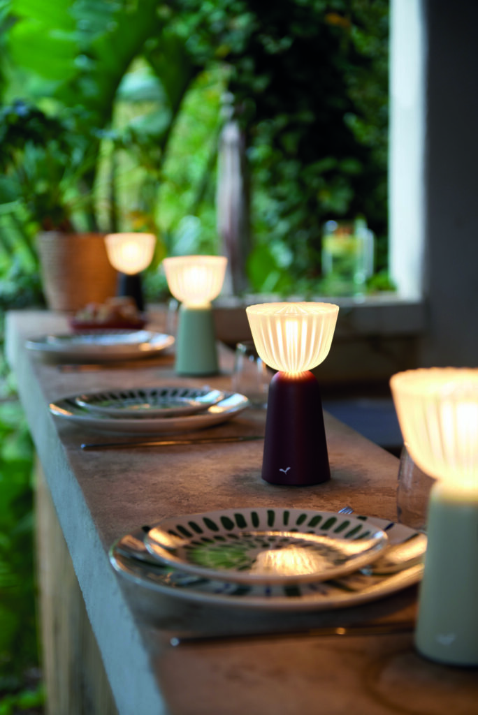 Une scène de repas en plein air éclairée par la Lampe Swiing 7 sur un rebord en pierre, complétée par des assiettes à motifs verts et un fond luxuriant et feuillu.