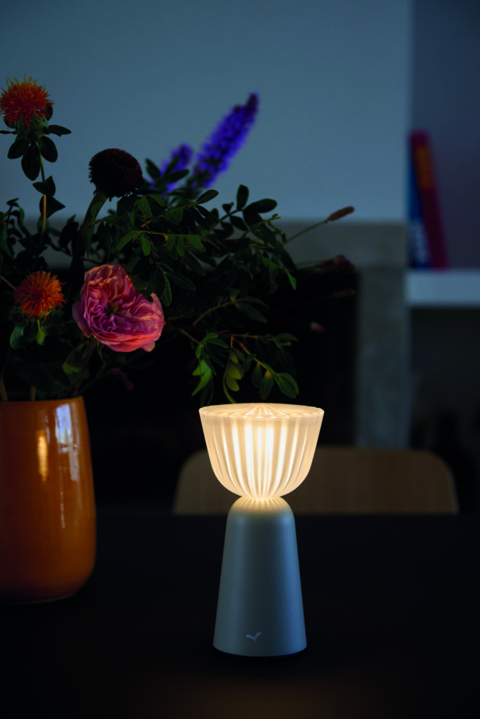 Une Lampe Swiing 7 moderne émet une douce lueur lorsqu'elle est posée sur une table à côté d'un vase rempli de fleurs colorées, telles que des roses roses et des fleurs orange.