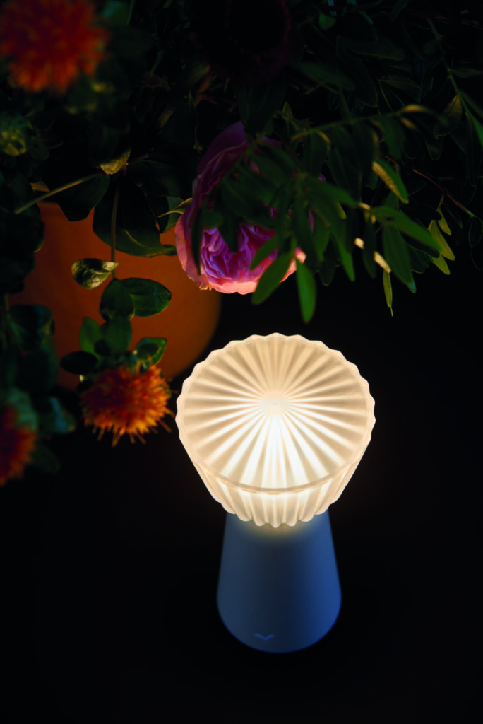 Une Lampe Swiing 7 lumineuse au design nervuré repose sur une surface sombre, illuminant doucement les fleurs et les feuilles à proximité.