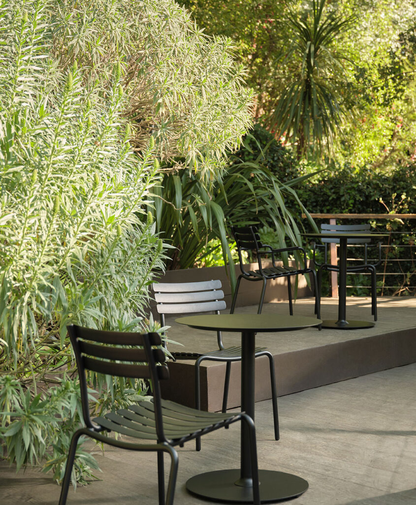 Le patio extérieur comprend des tables élégantes en métal noir et des chaises de salle à manger ALUA, entourées de plantes et d'arbres verts luxuriants.