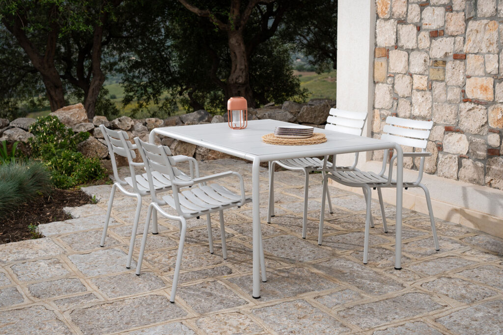 Dans un cadre paisible de patio extérieur, une table en métal blanc et quatre chaises de salle à manger ALUA avec accoudoirs reposent sur des dalles de pierre. Une pile d'assiettes et une lanterne orange ornent le plateau de la table, tandis que des arbres et un mur de pierre renforcent le charme naturel de cette scène paisible.