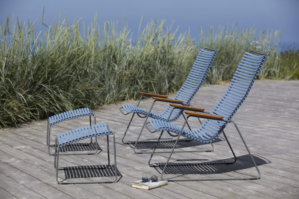 Trois chaises CLICK Lounge en bleu et métal ornent une terrasse en bois avec deux repose-pieds. De hautes herbes se balancent sous un ciel bleu paisible, tandis qu'un magazine et un livre sont posés sur la terrasse, parfaits pour se détendre.