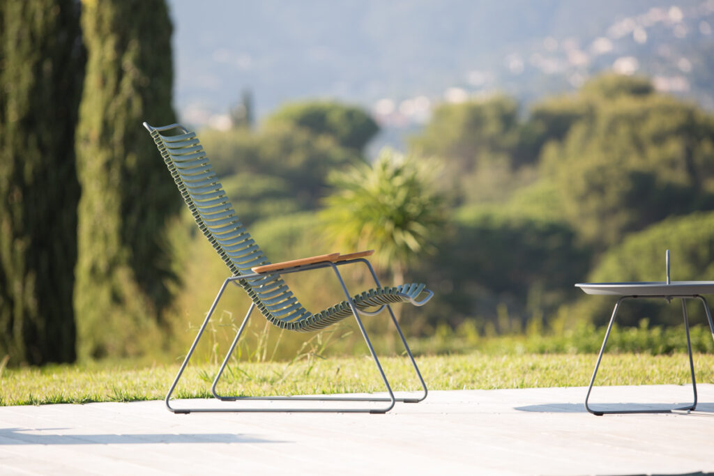 La chaise longue CLICK, dotée d'un design élégant en métal et d'accoudoirs en bois, se trouve sur une terrasse avec vue sur un paysage verdoyant et des collines lointaines.