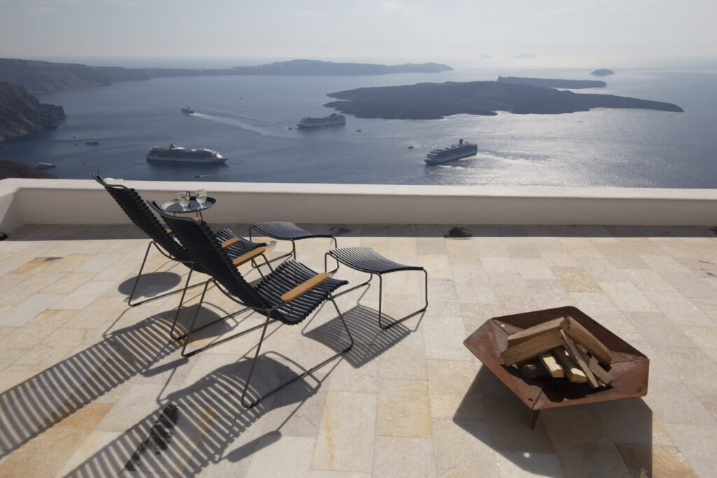Le patio comprend des chaises longues CLICK et une table avec une vue sereine sur le paysage marin, y compris les bateaux de croisière à l'horizon. Un foyer rempli de bois au premier plan est idéal pour des soirées agréables en bord de mer.