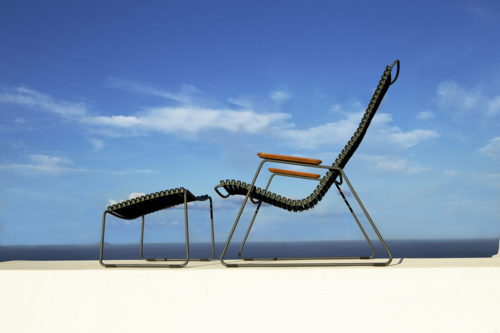 Un repose-pieds CLICK élégant complète une chaise longue moderne sur une surface blanche sous un ciel bleu parsemé de nuages.