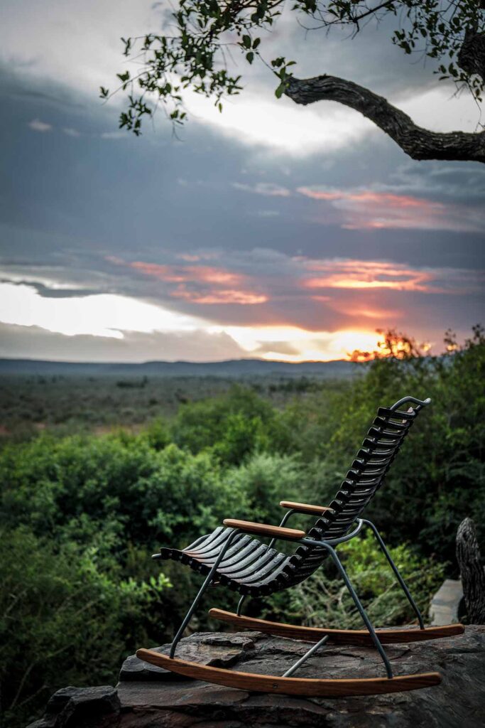 Une chaise à bascule pour enfants CLICK en noir est posée sur une terrasse en pierre, surplombant un paysage verdoyant tandis que le soleil se couche derrière des collines lointaines sous un ciel nuageux.