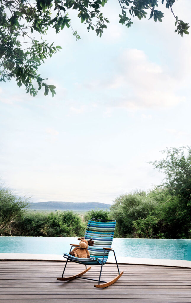 Un ours en peluche se détend sur la chaise à bascule pour enfants CLICK à rayures, située sur une terrasse en bois surplombant une piscine et une verdure luxuriante sous un ciel nuageux.