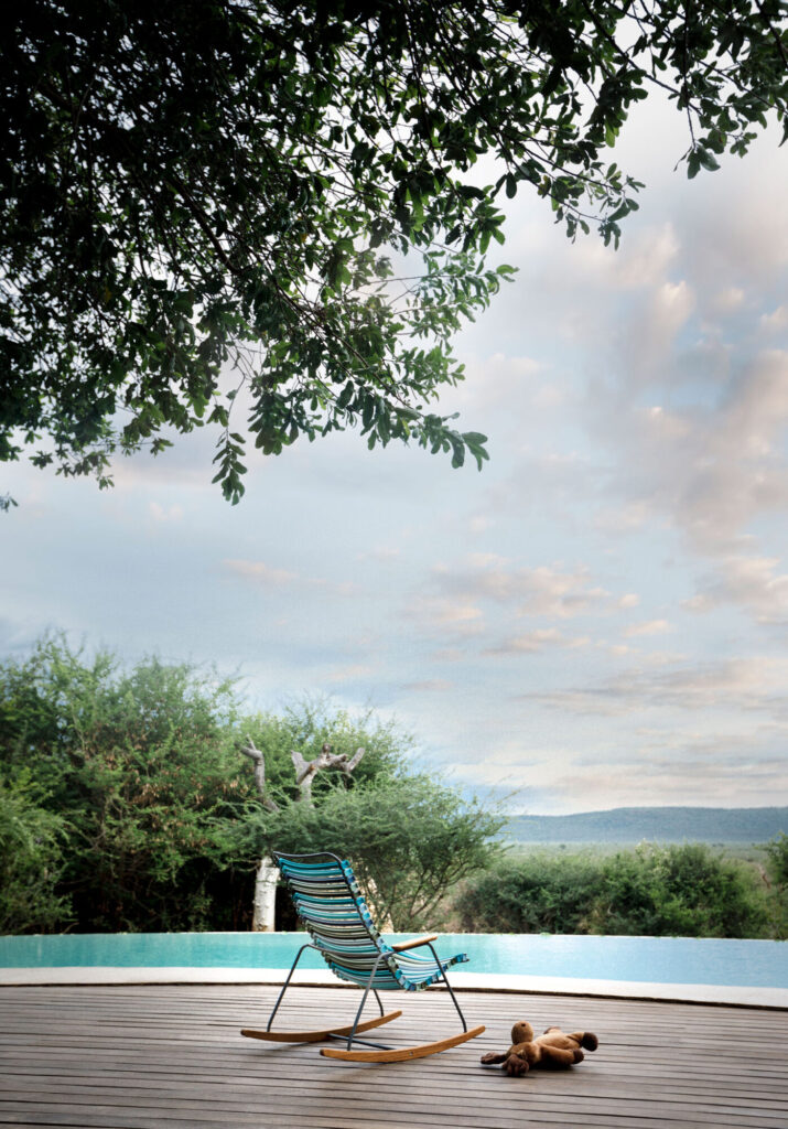 Une chaise à bascule et un ours en peluche pour enfants CLICK sont assis sur une terrasse en bois à côté d'une piscine extérieure, entourée d'arbres et d'un ciel nuageux, invitant à la tranquillité.
