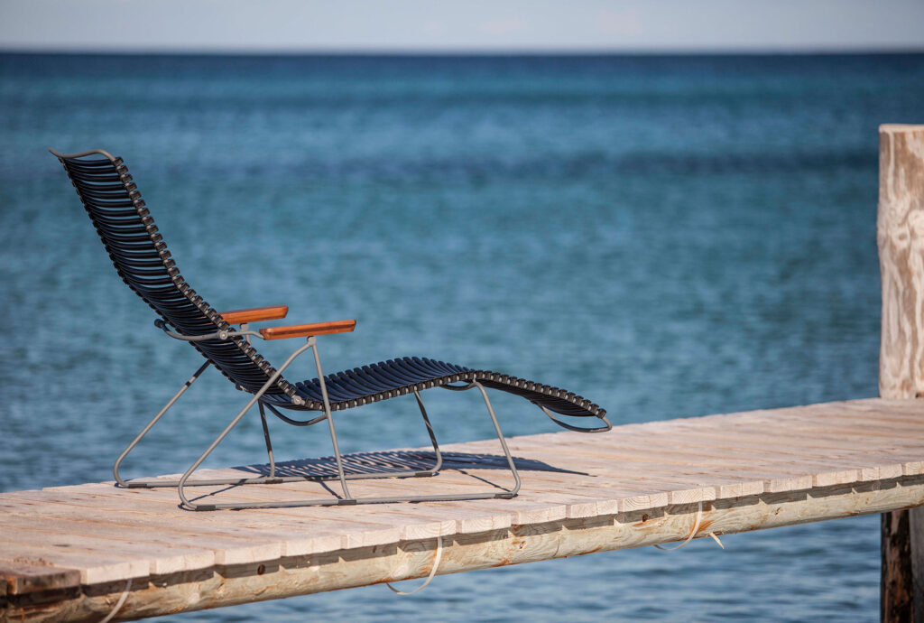 Une chaise longue CLICK noire avec accoudoirs en bois est installée sur une jetée en bois, offrant une vue sur la mer bleue tranquille sous un ciel clair.