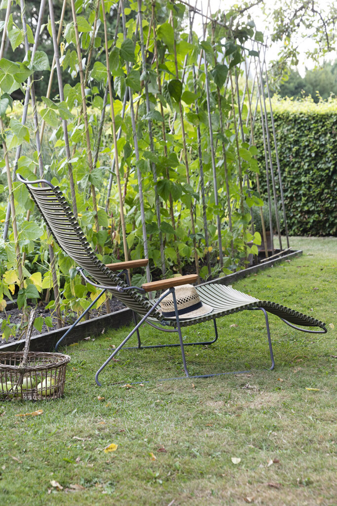 Le bain de soleil CLICK, aux teintes vertes et à la structure robuste en métal, repose élégamment sur l'herbe, accompagné d'un panier. En arrière-plan, une treille de jardin est ornée de plantes grimpantes.