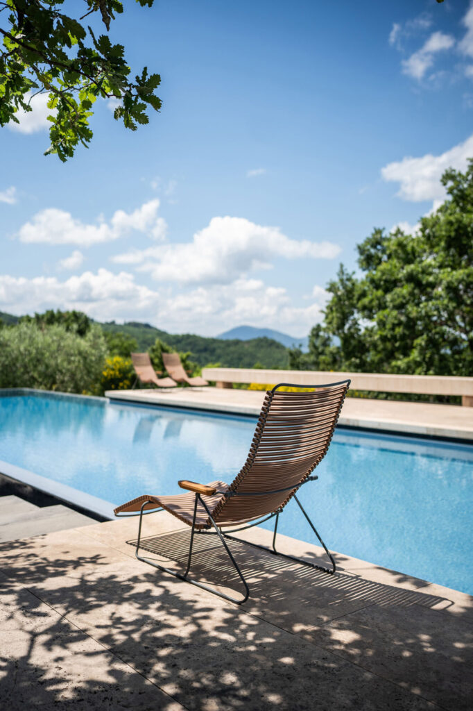 Le transat CLICK repose élégamment au bord d'une piscine scintillante sous un ciel partiellement nuageux, enveloppé d'arbres luxuriants et de collines lointaines.