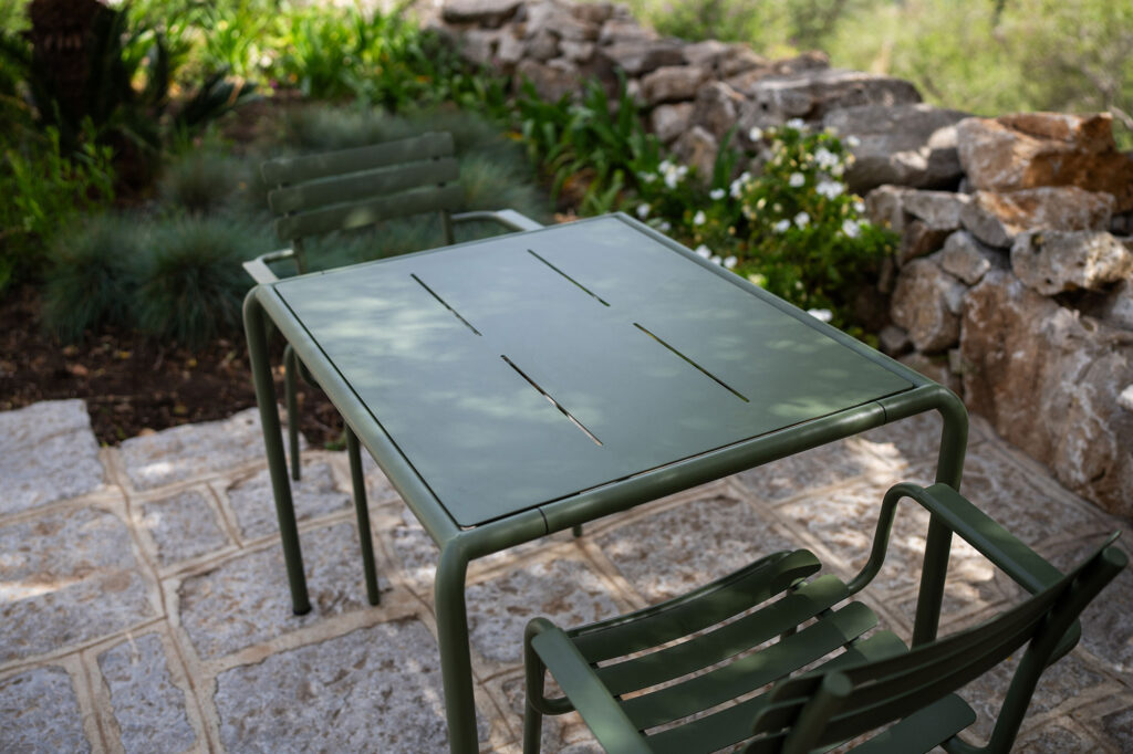 La table à manger en métal vert AVANTI, mesurant 83x98 cm, est associée à deux chaises sur un patio en pierre, entouré d'un mur de pierre et d'une verdure luxuriante.