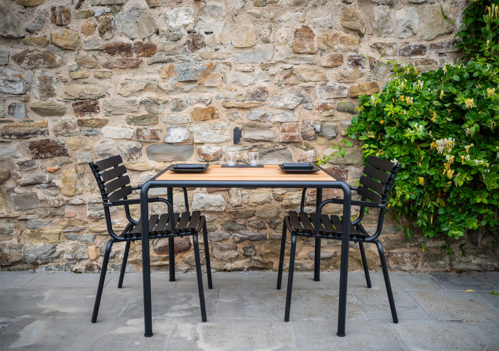 Deux chaises en métal noir et une table à manger AVANTI, mesurant 83x98 cm, sont posées contre un mur de pierre avec un feuillage vert sur le côté.