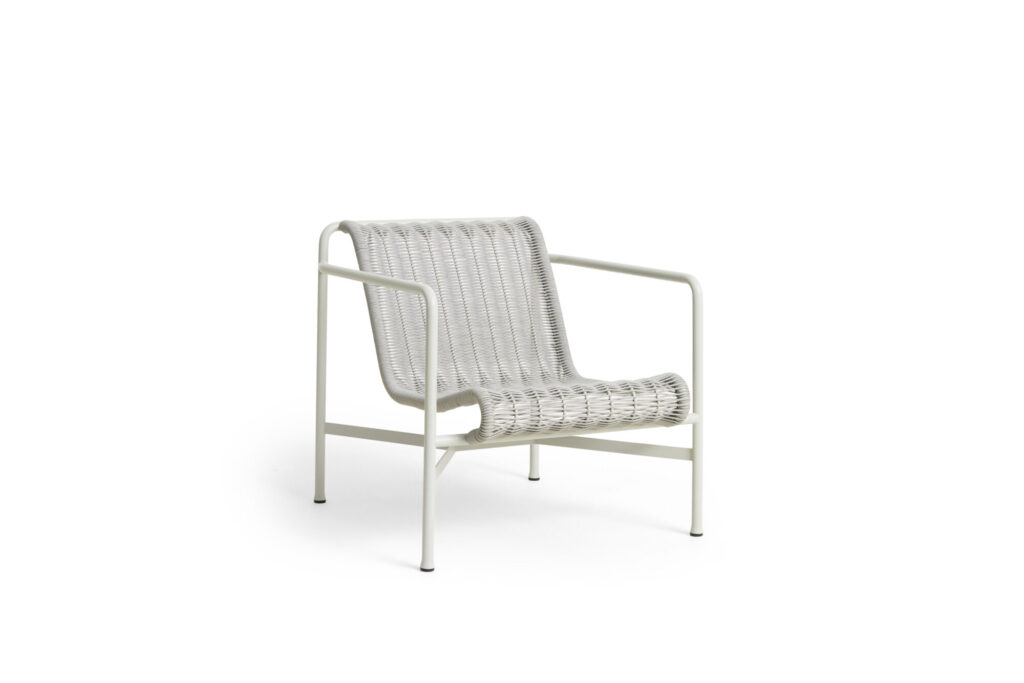 Une chaise longue tissée blanc crème avec une structure en métal minimaliste dans une nuance subtile de rouge fer, dotée d'une assise large et d'un dossier bas, sur un fond blanc sans ornements.