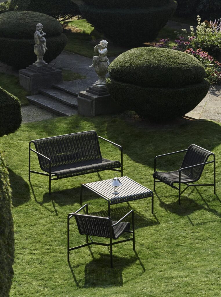 Ensemble de meubles de patio en métal comprenant deux chaises, une causeuse et une table en blanc crème élégant sur une pelouse, flanqué de buissons bien entretenus et de statues en pierre dans un cadre de jardin.