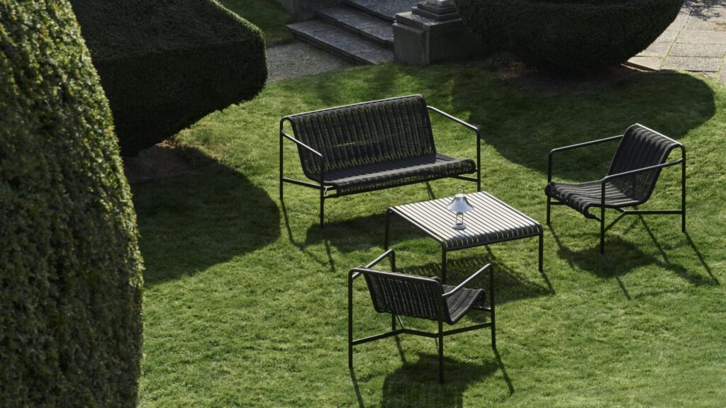 Un petit espace extérieur sur pelouse comprend un banc, deux chaises et une table carrée blanc crème. Entouré de haies taillées et d'un escalier en arrière-plan, c'est l'endroit idéal pour se détendre.