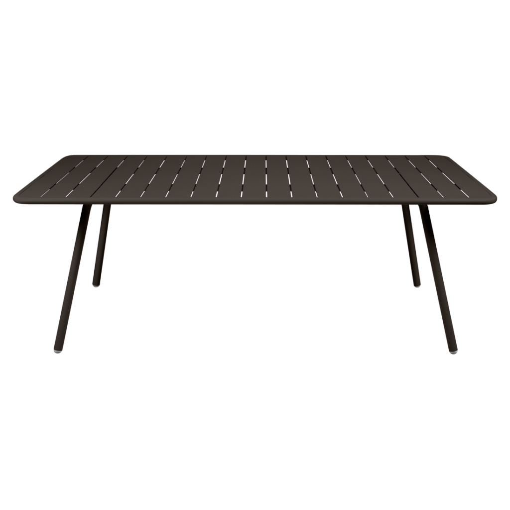 Une table rectangulaire marron foncé Luxembourg avec une surface à lattes et quatre pieds inclinés se dresse élégamment sur un fond violet clair uni, incarnant le mobilier d'extérieur moderne.