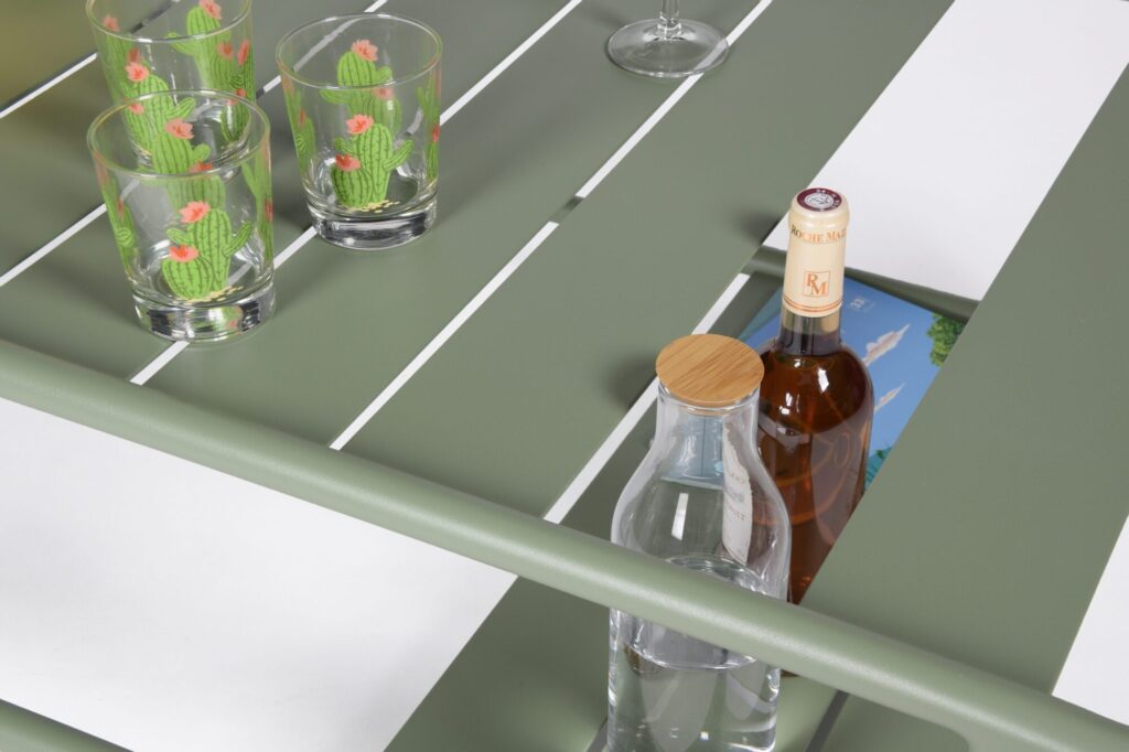 Une table basse verte avec ses verres trône au-dessus d'une piscine Maya d'un bleu serein. Quatre verres ornés de motifs de cactus, une bouteille d'eau, une bouteille de vin et un verre à vin vide sont disposés avec art sur les sections, ajoutant une touche de charme à ce décor pittoresque.