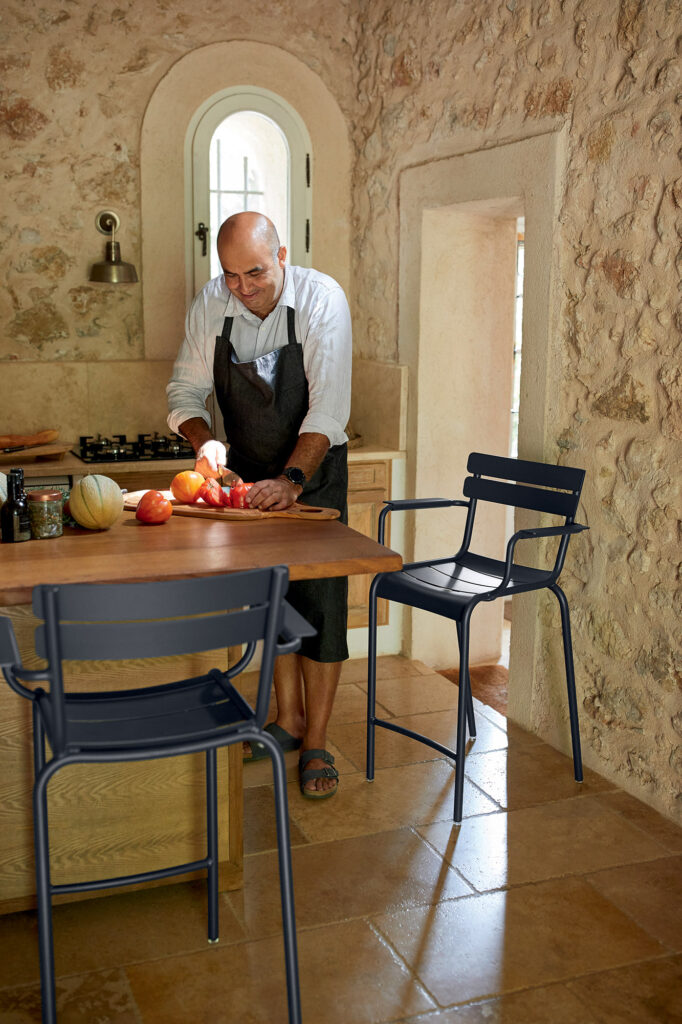 Un homme coupe des fruits sur un plan de travail en bois dans une cuisine luxembourgeoise pittoresque. Des murs en pierre et une petite fenêtre entourent la pièce, tandis que deux fauteuils trônent confortablement à la table.