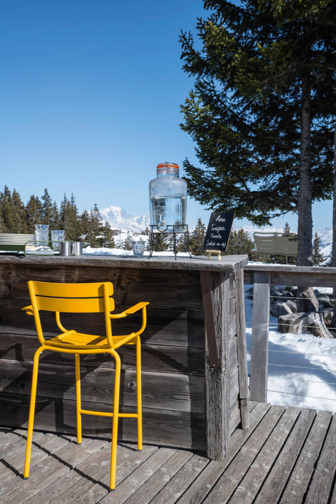 Un fauteuil jaune se trouve près d'un bar extérieur en bois surmonté d'un grand distributeur d'eau, situé dans le paysage enneigé du Luxembourg sous un ciel bleu clair.