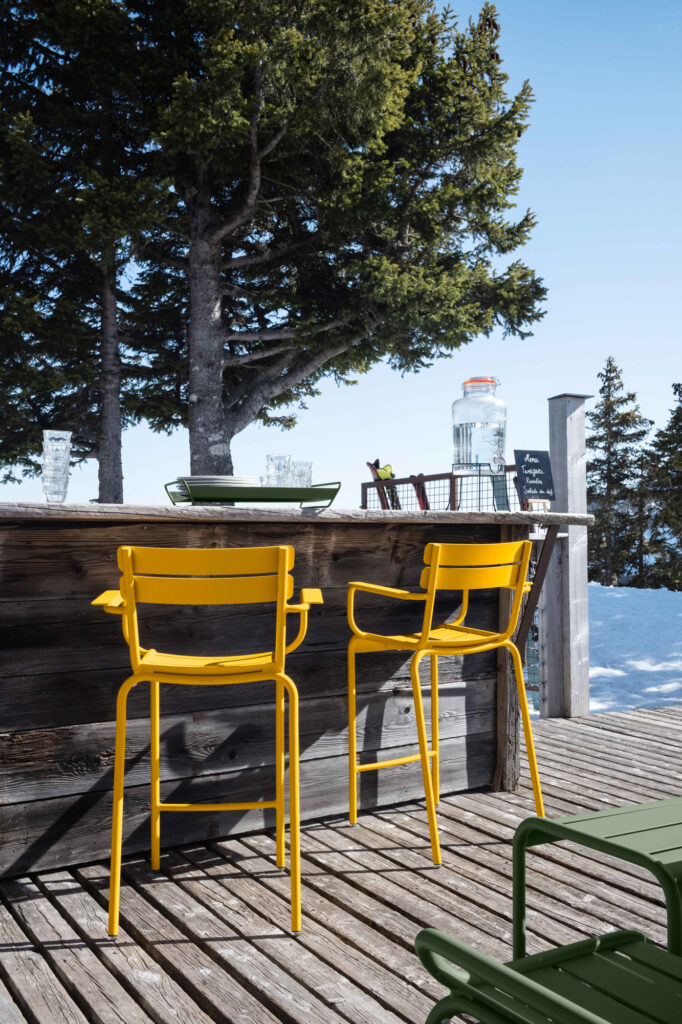 Deux fauteuils jaunes, installés sur une terrasse en bois, créent une ambiance chaleureuse et luxembourgeoise. Un grand bocal et des verres sont élégamment disposés sur le bar.