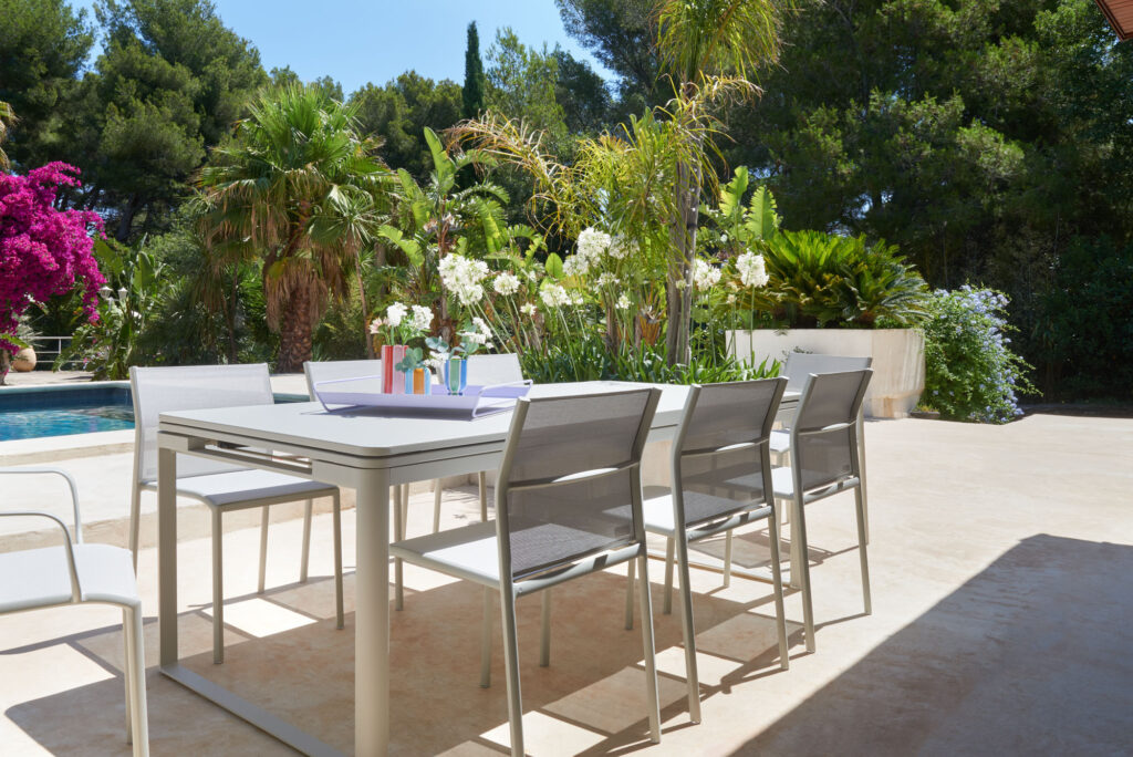 Espace repas extérieur avec table et chaises extensibles aux tons tonka sur une terrasse. La végétation environnante est composée d'arbres et de plantes. Une piscine est visible en arrière-plan par cette belle journée ensoleillée de Biarritz.