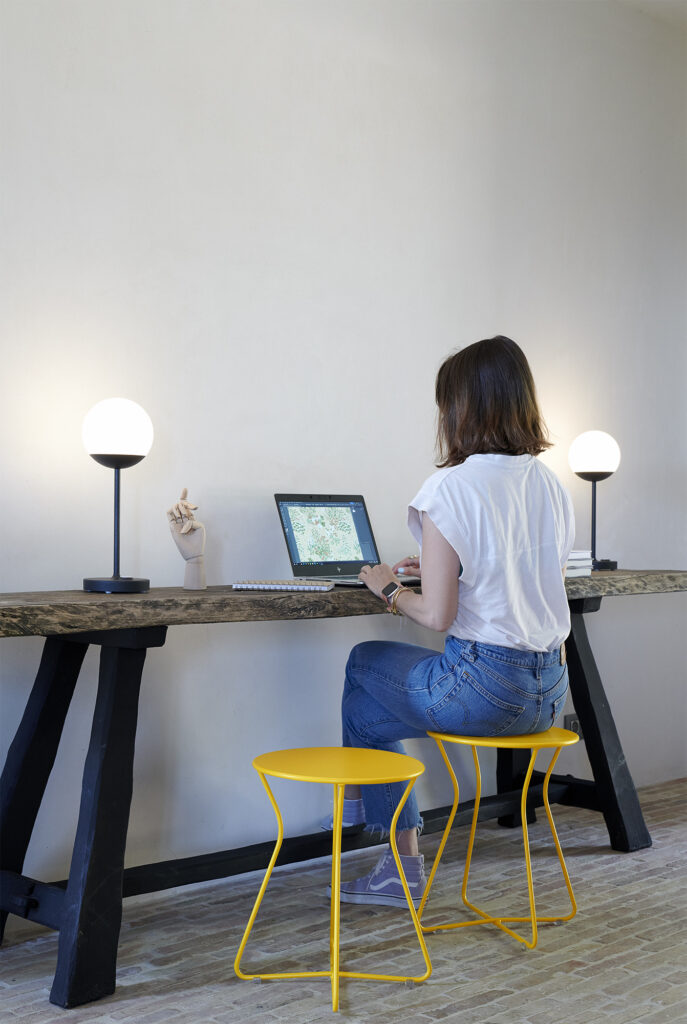 Une femme en chemise blanche et jean est assise sur un Tabouret Cocotte, devant un long bureau en bois, et utilise un ordinateur portable. Deux lampes sphériques et une maquette de main en bois sont également posées sur le bureau.