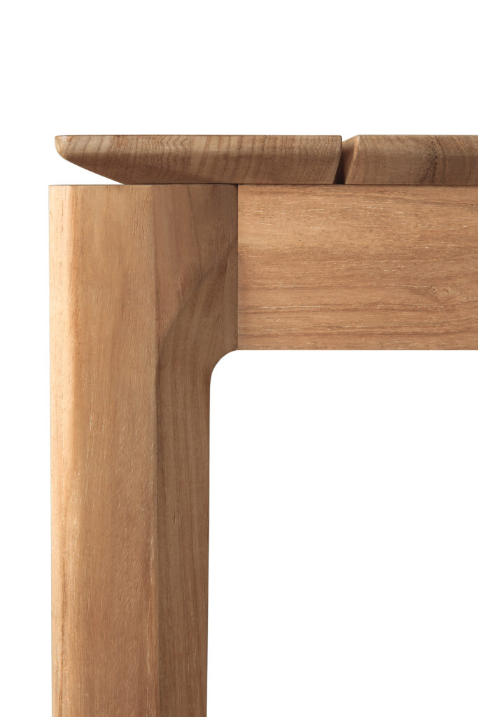 Gros plan d'un coin de table de jardin en teck avec une assiette partiellement posée sur le bord, sur fond blanc.