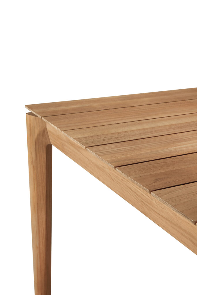 Vue rapprochée du coin d'une table de jardin en teck avec un plateau à lattes et des pieds inclinés, sur fond blanc.