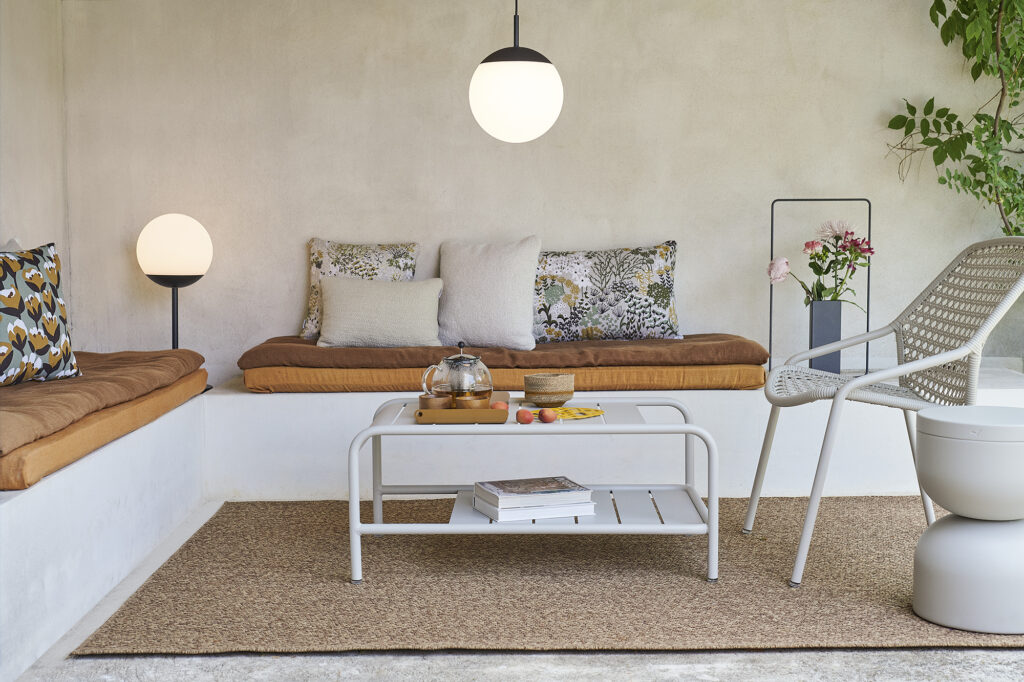 Espace salon extérieur avec bancs intégrés, coussins moelleux, une table basse Carbone contenant des articles de thé, une chaise, un lampadaire et une suspension, le tout posé sur un tapis tissé à côté d'une plante en pot luxuriante.