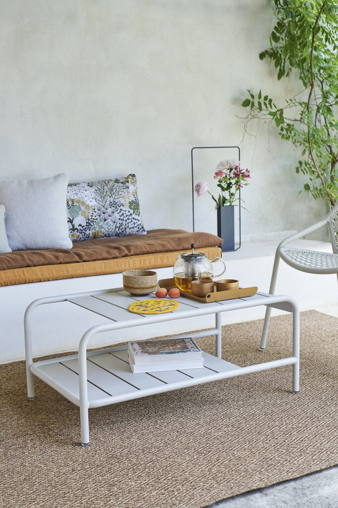 Une table basse blanche avec un service à thé et des collations repose sur un tapis tissé dans un coin salon confortable avec un banc rembourré, des oreillers, des plantes et le style signature Carbone.