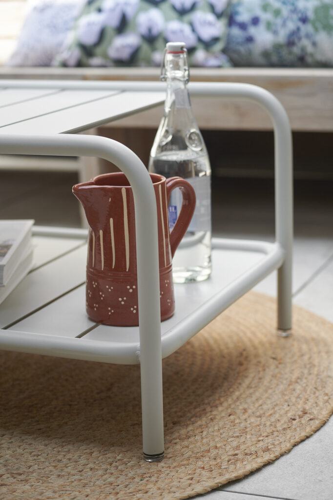 Un pichet en céramique rouge et une bouteille d'eau en verre sont posés sur l'étagère inférieure d'une table basse en métal blanc, posée sur un tapis rond tissé.