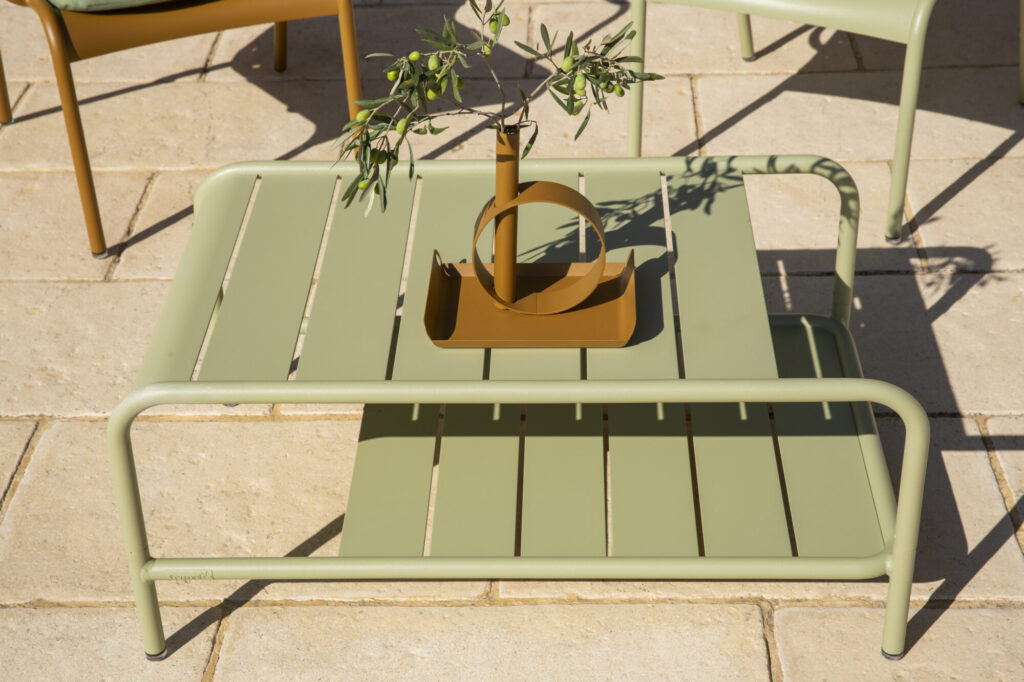 Une table basse Lxmbrg en métal vert avec un vase jaune moderne tenant une petite branche, posée sur un patio carrelé pour une touche inspirée du citron givré.