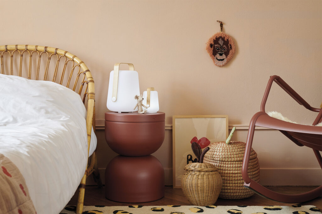 Un coin chambre soigneusement agencé avec un lit en rotin, deux tables d'appoint Tabouret empilées contenant des lampes blanches, des paniers tressés, une impression encadrée et une chaise berçante contre un mur beige.